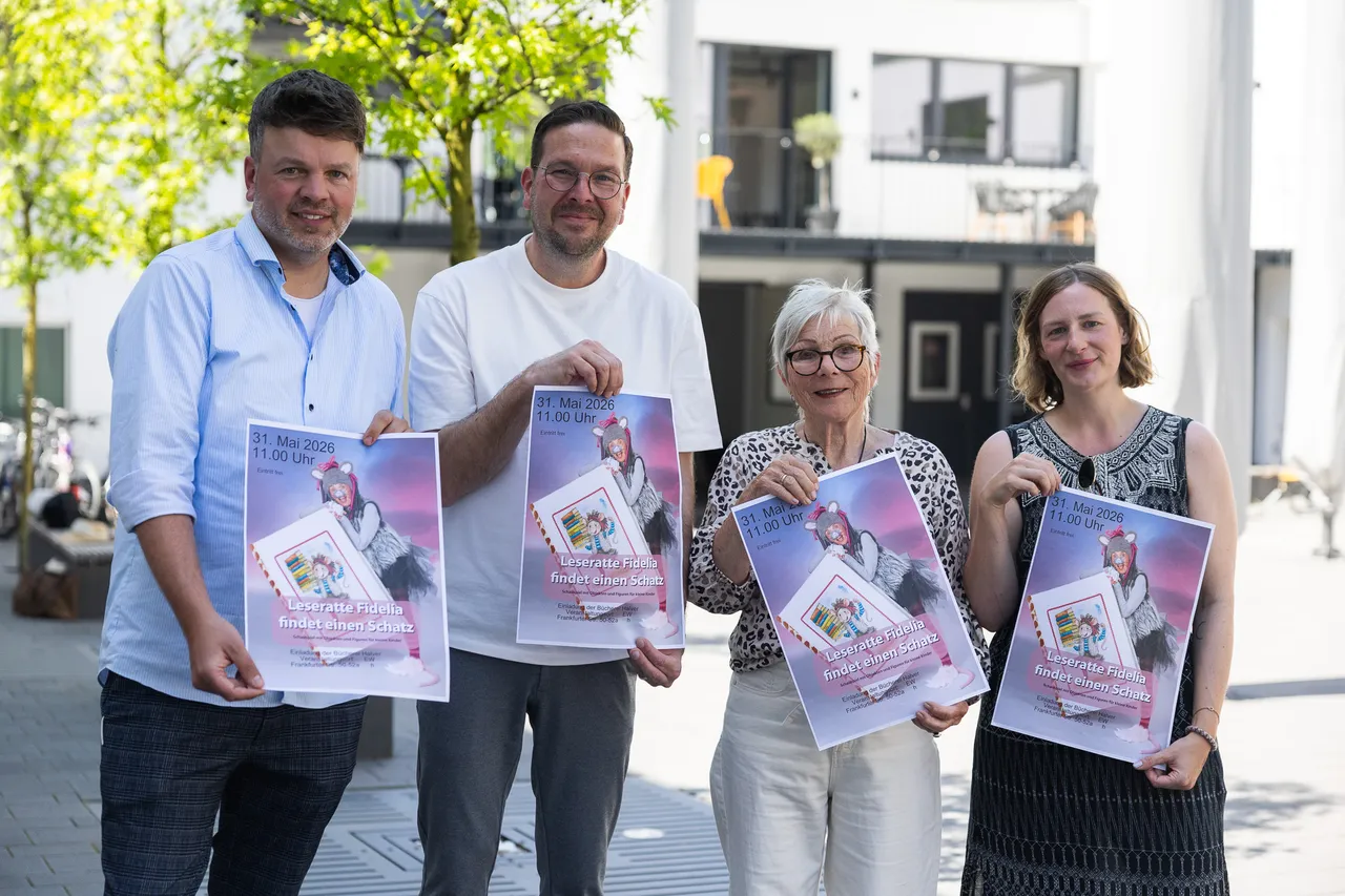 Vier Personen halten Plakate mit dem Titel „Leseratte Fidela findet einen Schatz“ in der Hand, vor dem Hintergrund eines hellen Gebäudes mit Säulen.