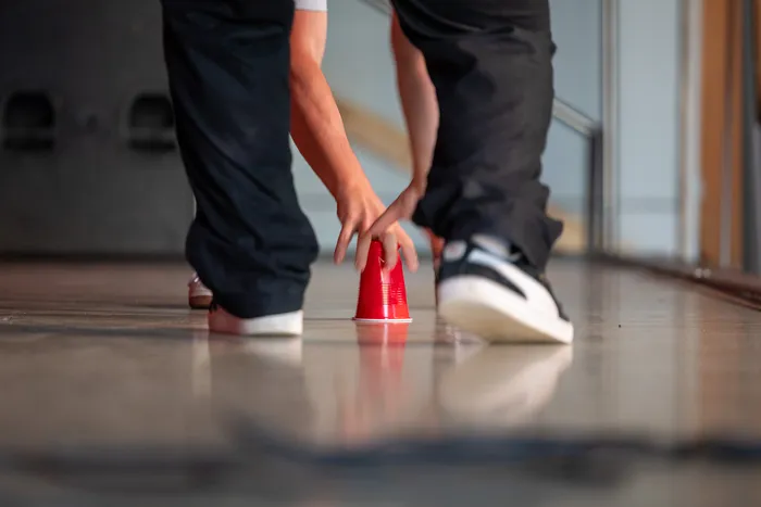 Zwei Personen stehen sich gegenüber und halten jeweils einen roten Plastikbecher in der Hand. Im Hintergrund ist eine Treppe und ein heller Raum zu sehen.