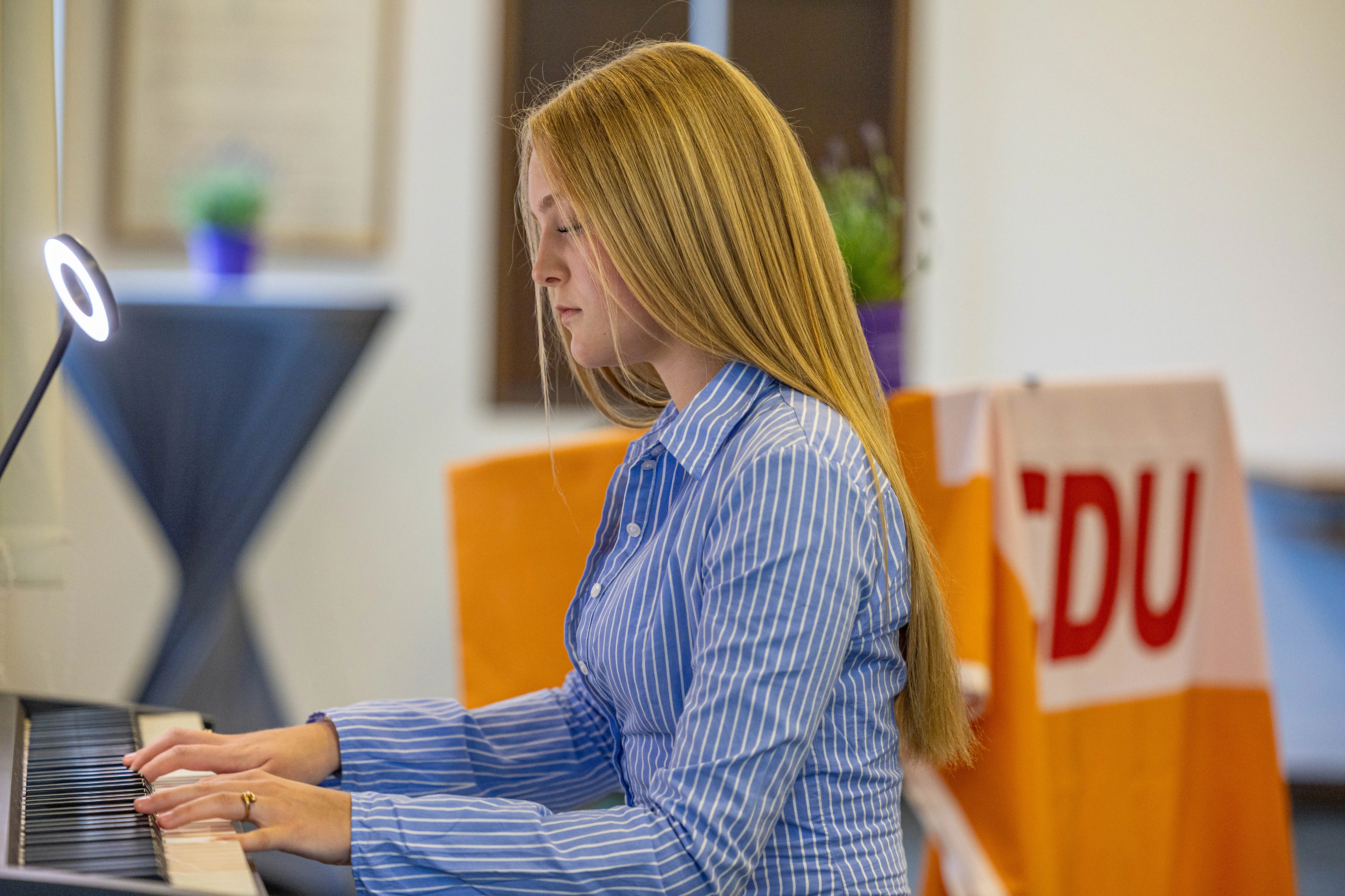 Eine junge Frau spielt auf einem Keyboard, während im Hintergrund ein Banner mit dem Kürzel „DU“ zu sehen ist.