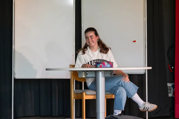 Eine junge Frau mit langen, lockigen Haaren sitzt lächelnd an einem Tisch auf einer Bühne. Sie trägt Jeans, ein weißes T-Shirt und Sneaker und hält eine Tasche in der Hand. Im Hintergrund eine weiße Leinwand und ein roter Vorhang.