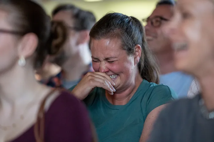 Eine Frau mit hochgestecktem Haar weint und bedeckt ihr Gesicht mit der Hand. Im Hintergrund sind weitere Menschen zu sehen.