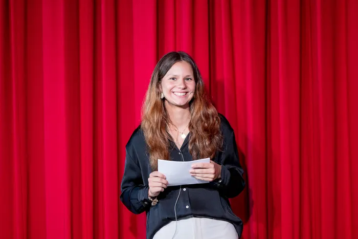 Eine junge Frau mit langen, gewellten Haaren lächelt in die Kamera, während sie ein Blatt Papier in der Hand hält. Im Hintergrund ein roter Vorhang.