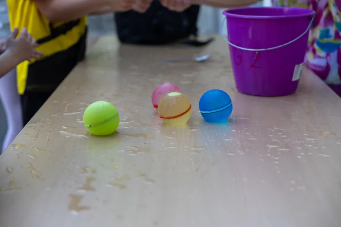 Drei Wasserbomben in den Farben Gelb, Rosa und Blau liegen auf einer nassen Holztischplatte. Im Hintergrund steht ein rosa Eimer und ein Teil einer Person mit gelbem T-Shirt ist zu sehen.