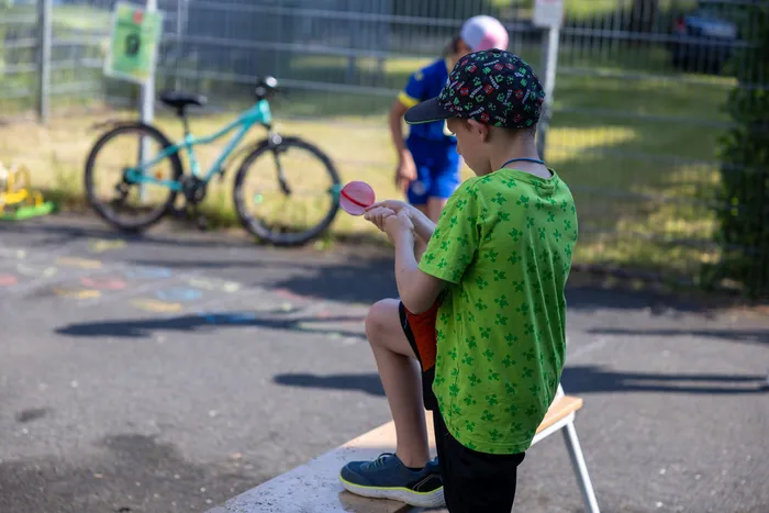 Ein Junge sitzt auf einer Bank und hält einen roten Kreisel in den Händen. Im Hintergrund steht ein blau-grünes Fahrrad und ein weiteres Kind.