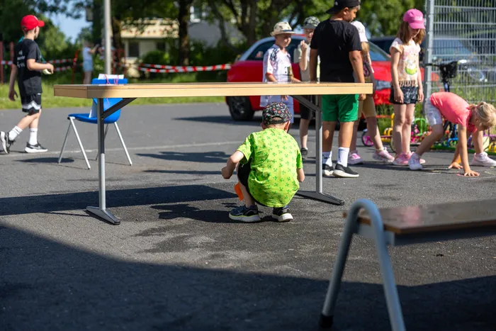 Ein Junge hockt auf dem Asphalt unter einem Tisch, während im Hintergrund Kinder spielen und Erwachsene stehen. Das Sportfest der Regenbogenschule findet im Freien statt.