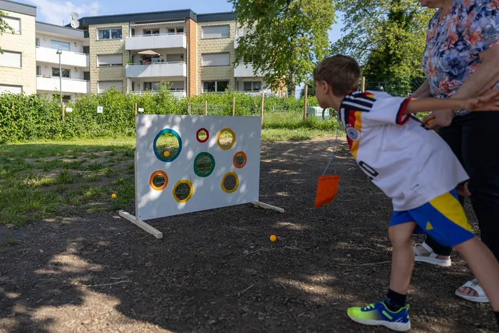 Ein Junge im Fußballtrikot der deutschen Nationalmannschaft wirft einen Ball auf eine selbstgebaute Zielscheibe mit unterschiedlich großen Löchern. Im Hintergrund ein Wohnhaus und Bäume.