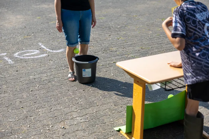 Eine Frau steht vor einem schwarzen Eimer, während ein Kind im Hintergrund einen Ball auf einen Tisch wirft. Im Hintergrund ist eine mit Kreide gezeichnete Ziellinie auf dem Asphalt zu sehen.
