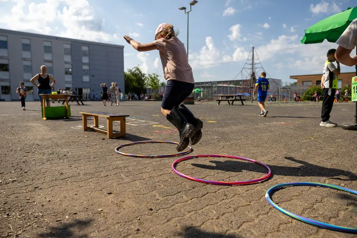 Ein Mädchen springt auf einem Schulhof über bunte Hula-Hoop-Reifen. Im Hintergrund sind weitere Kinder und ein Schulgebäude zu sehen.