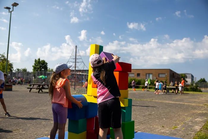 Zwei Kinder bauen auf einem Schulhof einen Turm aus bunten Schaumstoffblöcken. Im Hintergrund sind weitere Kinder und ein Spielplatz zu sehen.
