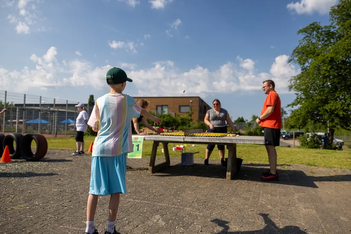 Ein Junge mit grüner Kappe und Startnummer 1 wirft einen Ball auf eine Tischtennisplatte, während eine Frau im Hintergrund steht und ein Mann zusieht. Im Hintergrund sind weitere Personen und ein Sportplatz zu sehen.