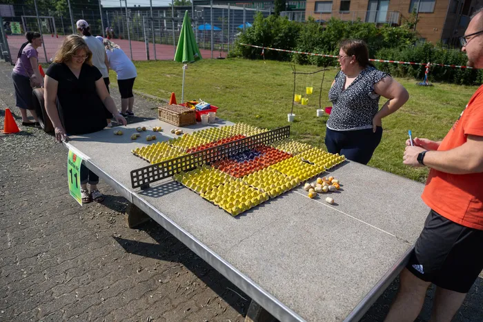 Ein Spielbrett mit vielen gelben und einigen roten/orangen Eiern steht auf einem Tisch im Freien. Menschen stehen um den Tisch und beobachten oder beteiligen sich am Spiel. Im Hintergrund ein Sportplatz und Gebäude.