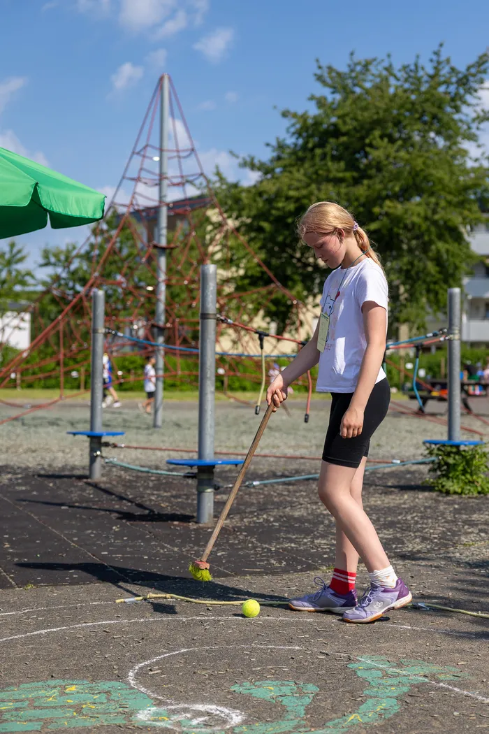 Ein Mädchen mit blonden Zöpfen spielt auf einem Spielplatz mit einem Besen und einem Ball. Im Hintergrund ist ein rotes Klettergerüst und ein grüner Sonnenschirm zu sehen.