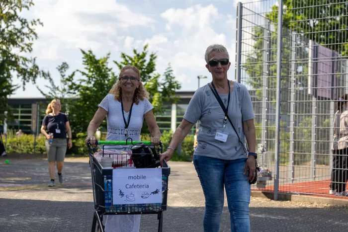 Zwei Frauen schieben einen mit Getränken und Gebäck gefüllten mobilen Cafeteria-Wagen über einen Platz. Im Hintergrund sind weitere Menschen und ein Zaun zu sehen.