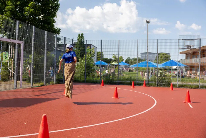 Eine Person rennt auf einer roten Tartanbahn zwischen orangefarbenen Hütchen. Im Hintergrund ein Basketballfeld, ein Zaun und Menschen bei einem Sportfest.