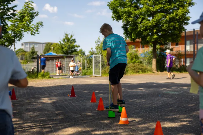 Ein Junge springt auf einem Sportplatz über ein gelbes Gummiseil, das zwischen orangefarbenen Hütchen gespannt ist. Im Hintergrund sind weitere Kinder und Gebäude zu sehen.