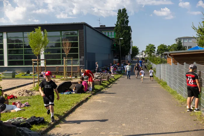 Kinder und Jugendliche bewegen sich auf einem Weg zwischen einem Schulgebäude und einem Spielplatz. Einige liegen oder sitzen im Schatten von Bäumen, andere laufen oder stehen in Gruppen. Im Hintergrund sind weitere Gebäude und Bäume zu sehen.