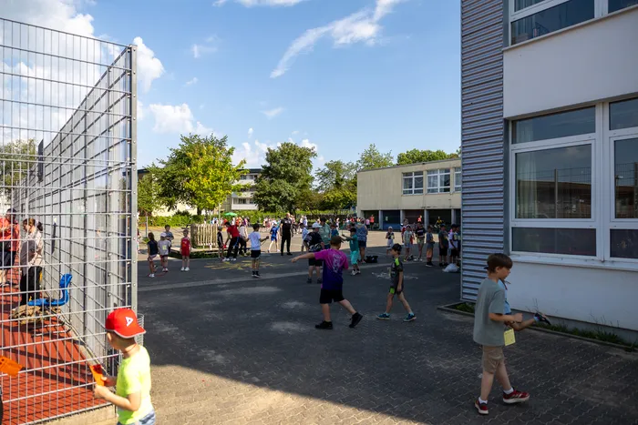 Kinder spielen und toben auf dem Schulhof der Regenbogenschule während eines Sportfestes an einem sonnigen Tag. Im Hintergrund sind Gebäude und Bäume zu sehen.