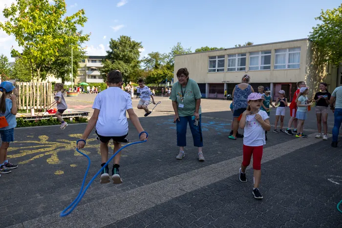 Ein Junge springt mit einem blauen Seil auf einem Schulhof, während eine Frau im Hintergrund zusieht. Weitere Kinder und Erwachsene sind auf dem Hof verteilt.