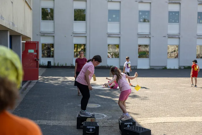 Zwei Mädchen stehen auf schwarzen Kisten und halten gelbe Bänder fest, während sie an einem Sportfest teilnehmen. Im Hintergrund steht ein Schulgebäude und weitere Kinder sind zu sehen.