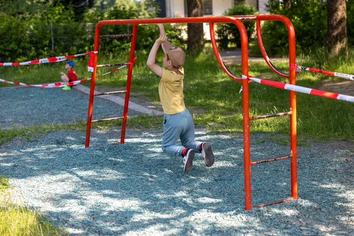 Ein Kind hängt an einer roten Reckstange auf einem blauen Spielplatz. Im Hintergrund ist eine abgesperrte Fläche mit rotem Absperrband zu sehen.