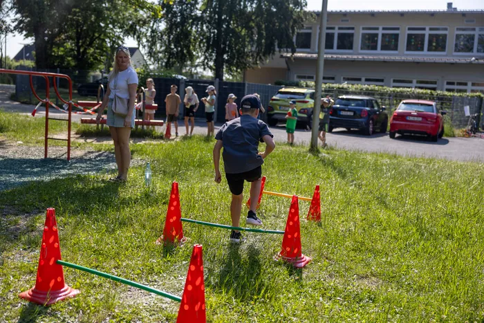 Ein Junge mit Baseballkappe überwindet bei einem Sportfest eine Hürdenreihe aus orangefarbenen Pylonen auf einer grünen Wiese. Im Hintergrund stehen weitere Kinder und eine erwachsene Frau sowie ein Schulgebäude und parkende Autos.