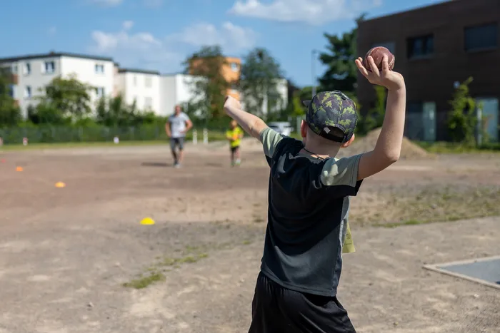 Ein Junge wirft einen American-Football auf einem Sportplatz, während im Hintergrund andere Personen trainieren. Im Hintergrund sind Wohnhäuser und Bäume zu sehen.