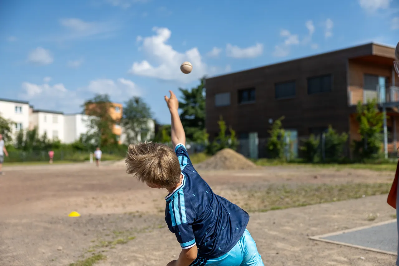 Ein Junge wirft einen Ball in die Luft. Im Hintergrund sind Gebäude und weitere Personen auf einem Sportplatz zu sehen.