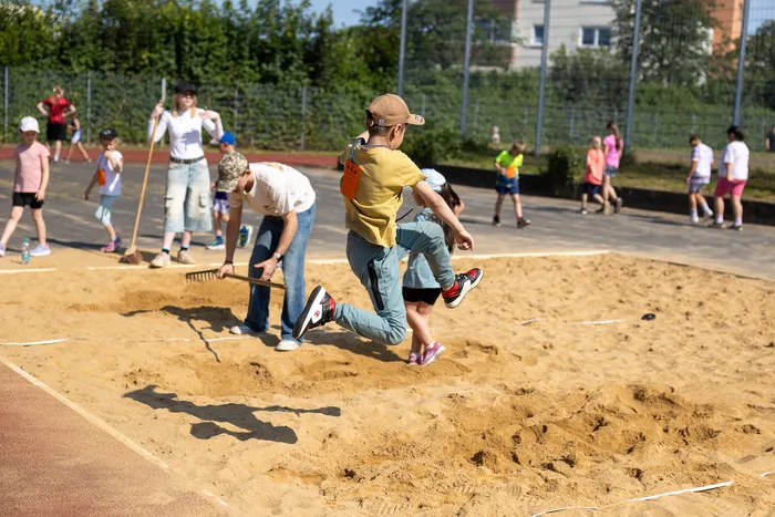 Ein Schüler springt in der Sandgrube während eines Sportfestes. Im Hintergrund sind weitere Kinder und Erwachsene zu sehen, die bei der Vorbereitung helfen.