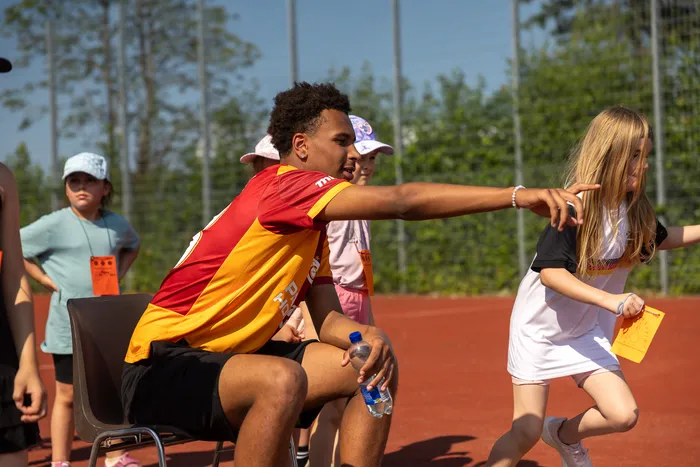 Ein junger Mann mit dunkler Hautfarbe zeigt mit ausgestrecktem Arm nach rechts, während er auf einem Stuhl sitzt. Im Hintergrund stehen und bewegen sich Kinder und Jugendliche auf einem roten Tennisplatz.