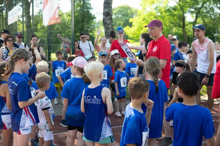 Eine Gruppe von Kindern in Sportkleidung steht auf einem Sportplatz und lauscht den Anweisungen eines Mannes mit einer roten Kappe und einer Zahl 6 in der Hand. Im Hintergrund sind weitere Zuschauer und ein Baum zu sehen.
