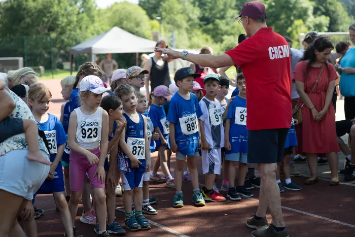 Ein Mann mit rotem Shirt und Baseballkappe zeigt mit ausgestrecktem Arm nach links auf eine Gruppe von Kindern in Laufkleidung mit Startnummern. Im Hintergrund sind weitere Zuschauer und Zelte zu sehen.