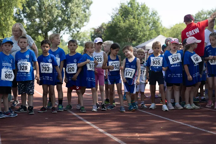 Eine Gruppe von Kindern steht auf einer roten Laufbahn bereit für den Start eines Cross- und Querlaufrennens. Die Kinder tragen blaue Trikots mit Startnummern und werden von erwachsenen Betreuern und Zuschauern umgeben.