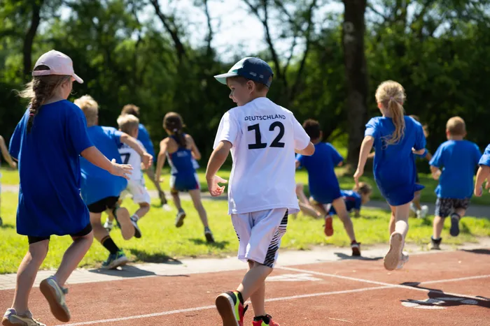 Eine Gruppe Kinder rennt auf einer roten Tartanbahn, wobei ein Junge im weißen Trikot mit der Nummer 12 und dem Aufdruck 