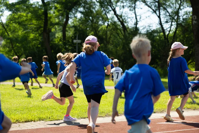 Eine Gruppe Kinder rennt bei einer Sportveranstaltung im Freien auf einer roten Tartanbahn. Viele tragen blaue T-Shirts und einige haben rosa Kappen. Bäume sind im Hintergrund zu sehen.