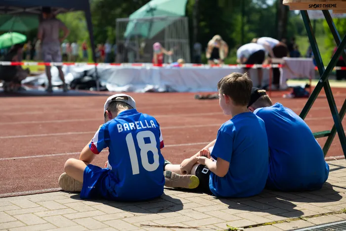 Drei junge Sportler in blauen Trikots mit den Nummern 18 und weiteren, sitzen auf dem Boden neben einer roten Laufbahn. Im Hintergrund sind weitere Läufer und Zuschauer zu sehen.