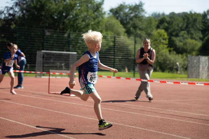 Ein blondhaariger Junge in Sportkleidung läuft auf einer roten Tartanbahn, während eine Frau im Hintergrund applaudiert. Weitere Läufer sind im Hintergrund zu sehen.