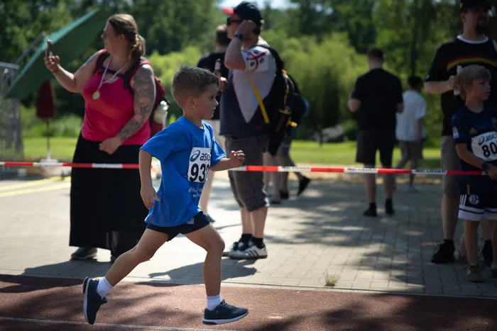 Ein Junge mit der Startnummer 93 rennt bei einem Crosslauf über eine niedrige Hürde. Im Hintergrund sind weitere Läufer und Zuschauer zu sehen.