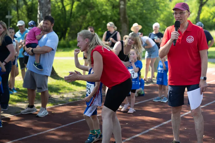 Eine Frau in roter Sportkleidung motiviert ein Kind in Laufkleidung auf einer roten Tartanbahn. Im Hintergrund stehen und sitzen Zuschauer, während ein Mann mit Mikrofon die Veranstaltung moderiert.