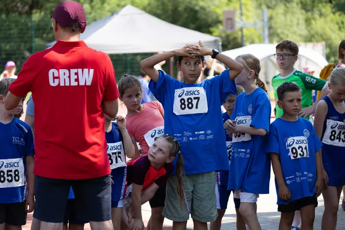 Eine Gruppe von Kindern in blauen T-Shirts mit Startnummern steht aufgereiht und blickt erwartungsvoll vor sich. Ein Betreuer in rotem Shirt mit der Aufschrift 
