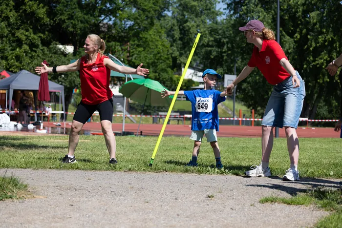 Zwei Frauen unterstützen einen Jungen beim Stabhochsprung auf einer Sportanlage. Die Athleten tragen Sportkleidung und Startnummern.