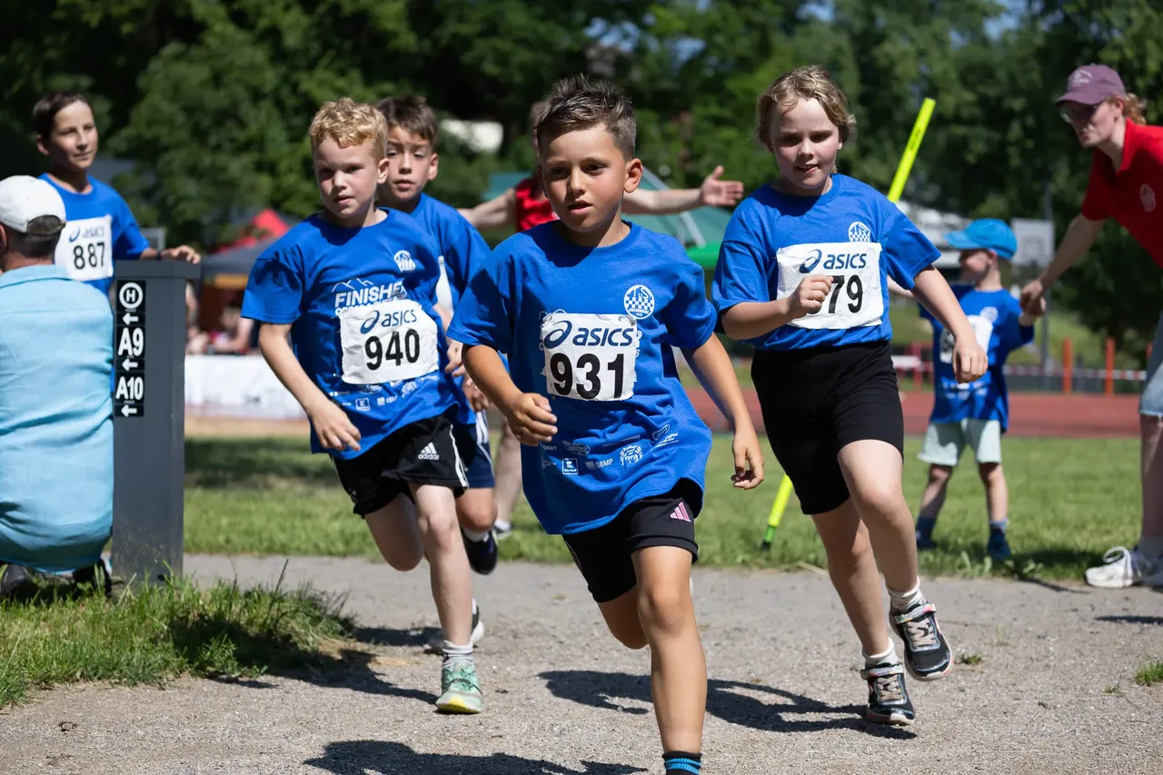 Eine Gruppe von Kindern rennt bei einem Crosslauf. Die Läufer tragen blaue T-Shirts mit Startnummern und Sportkleidung.