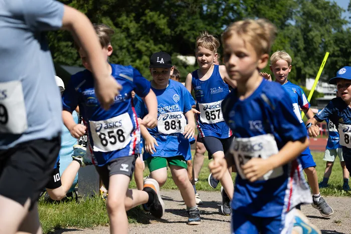 Eine Gruppe von Kindern rennt bei einem Crosslauf. Die Läufer tragen Startnummern und Sportkleidung, einige haben Mützen auf.