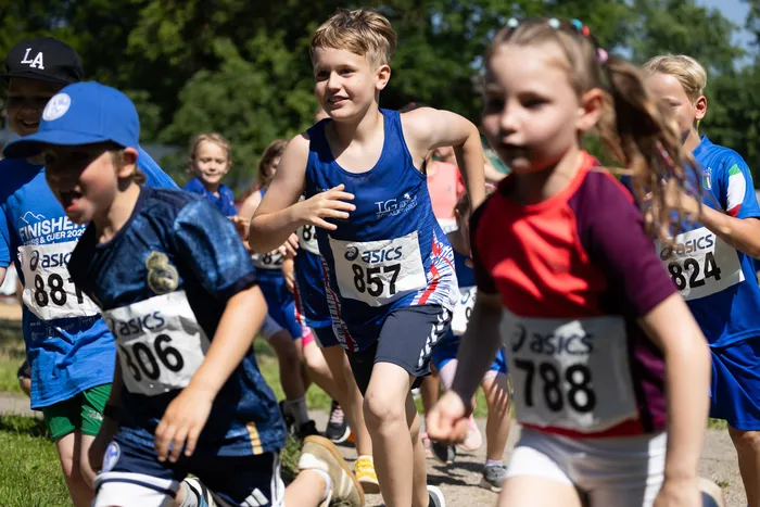 Eine Gruppe Kinder rennt bei dem Cross- und Querlauf 2026 mit Startnummern auf der Brust. Der Junge im Vordergrund trägt ein blaues Sportshirt und lächelt in die Kamera.