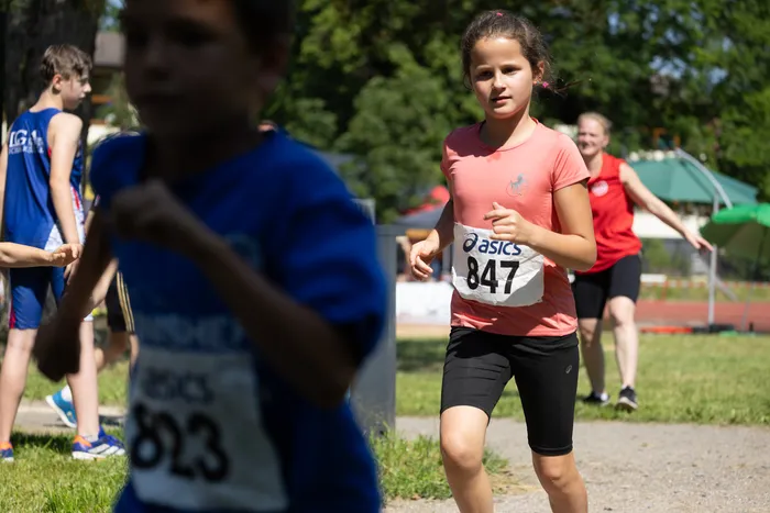 Ein Mädchen mit der Startnummer 847 läuft bei einem Crosslauf. Im Hintergrund sind weitere Läufer und Zuschauer zu sehen.