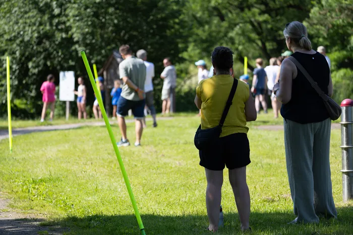 Menschen stehen und gehen auf einer Wiese, einige mit gelben Stäben markiert. Im Vordergrund fotografiert eine Person mit gelbem Shirt die Szene.