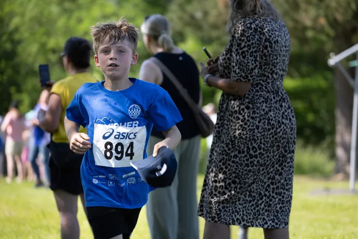 Ein Junge mit der Startnummer 894 läuft bei einem Crosslauf. Im Hintergrund steht eine Frau und filmt ihn mit einem Smartphone.