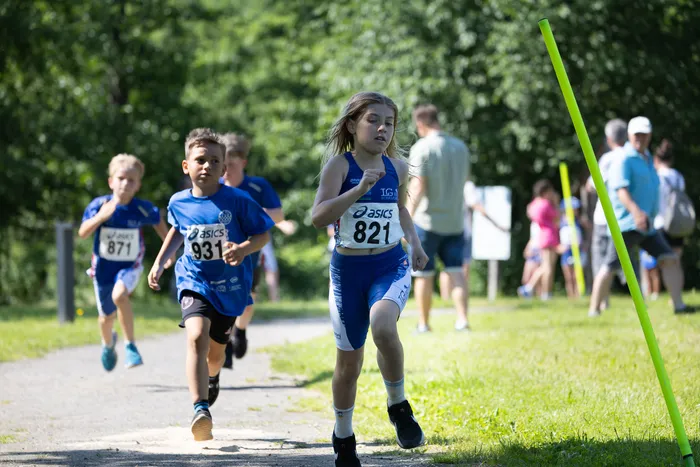 Eine Gruppe von Kindern rennt bei einem Crosslauf auf einem Pfad. Im Vordergrund trägt ein Mädchen mit der Startnummer 821 ein blaues Laufoutfit und konzentriert sich auf das Rennen. Im Hintergrund sind weitere Läufer und Zuschauer zu sehen.