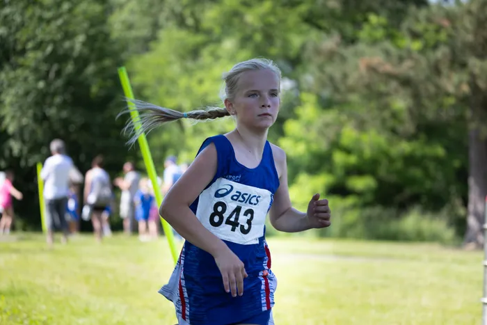 Ein junges Mädchen mit blonden Zöpfen läuft bei einem Crosslauf, trägt eine Startnummer (843) und ein blaues Sporttrikot mit dem ASICS-Logo. Im Hintergrund sind weitere Läufer und Zuschauer auf einer Wiese zu sehen.