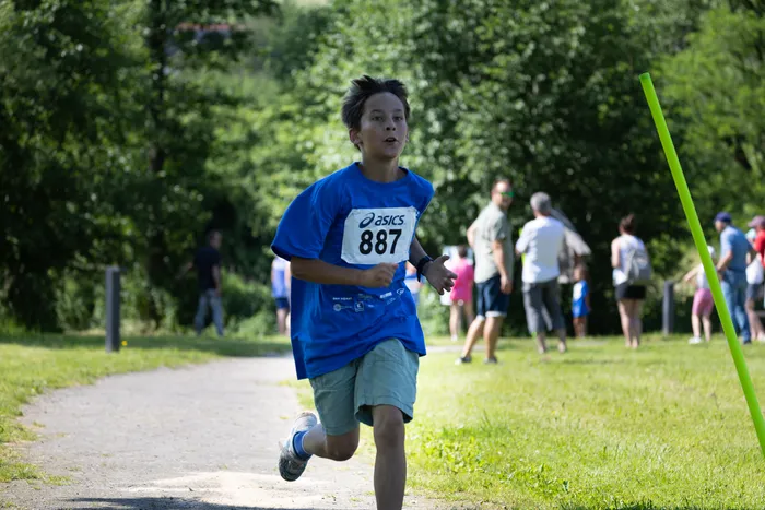 Ein Junge mit der Startnummer 887 läuft bei einem Crosslauf auf einer Waldweg. Im Hintergrund sind weitere Läufer und Zuschauer zu sehen.