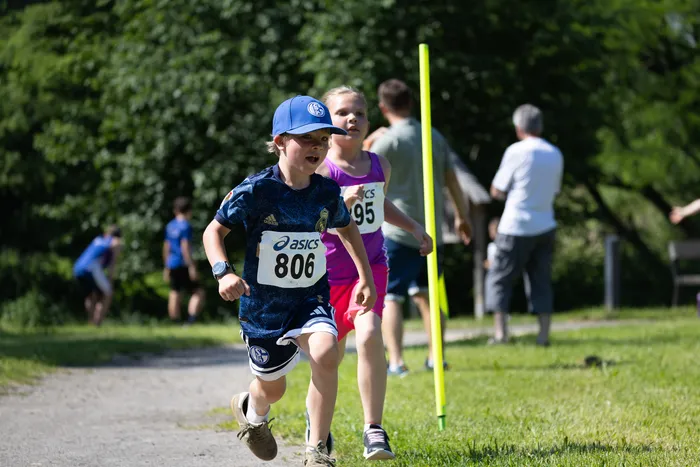 Ein Junge mit der Startnummer 806 rennt bei einem Crosslauf vor einem Mädchen mit der Startnummer 95. Im Hintergrund sind weitere Läufer und Markierungshütchen zu sehen.